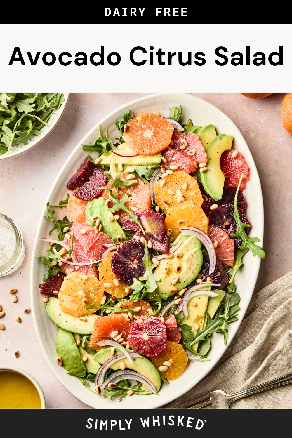 Oval plate of avocado citrus salad with arugula, grapefruit, blood orange, orange slices, avocado, red onion, and pine nuts. A fork, towel, and bowl of arugula are nearby. Text: Dairy Free Avocado Citrus Salad—a vibrant and refreshing avocado citrus salad perfect for any occasion.