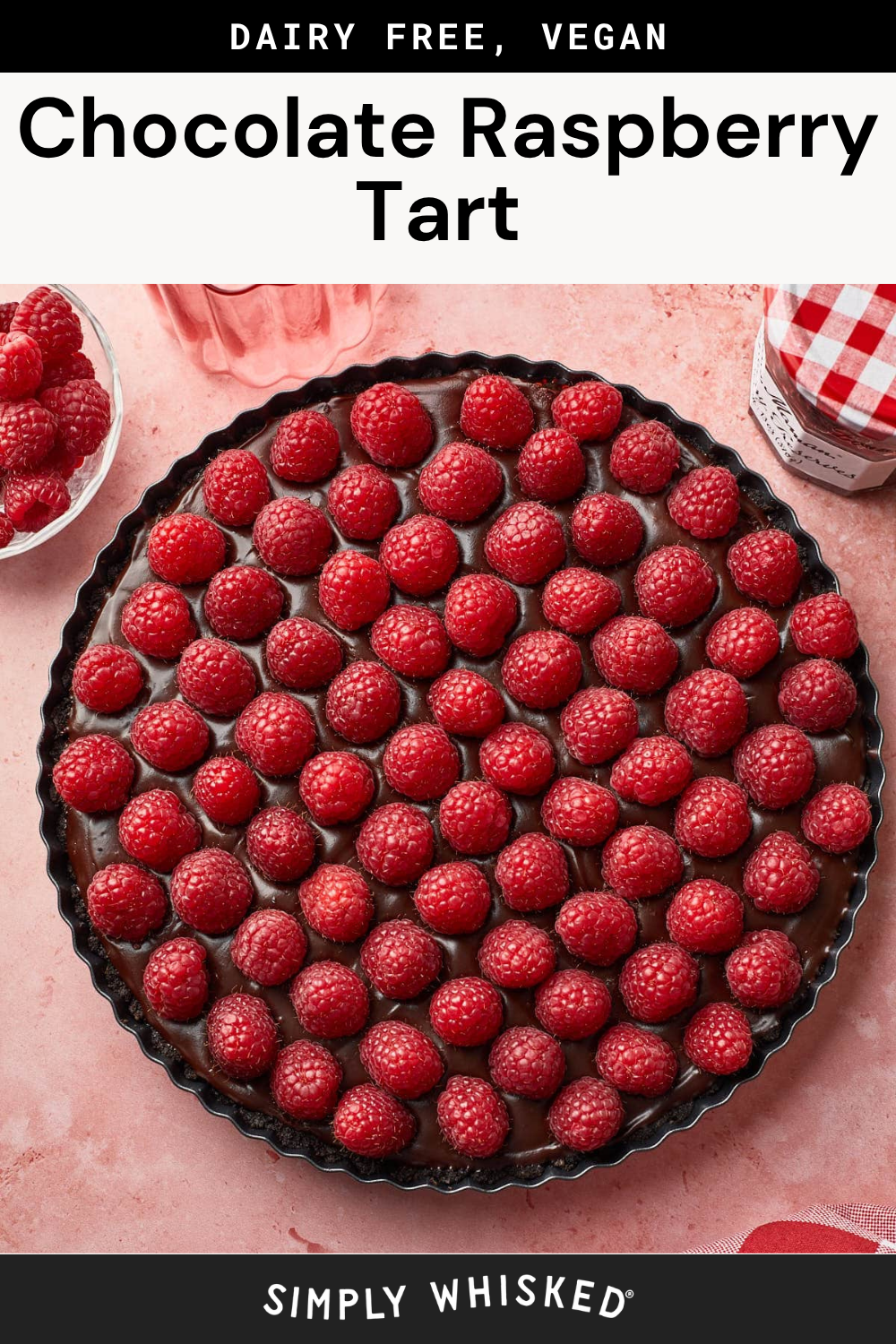 A chocolate raspberry tart topped with fresh raspberries sits on a pink surface, surrounded by a jar of jam, a glass of raspberries, and loose raspberries. Text reads Dairy Free, Vegan Chocolate Raspberry Tart.