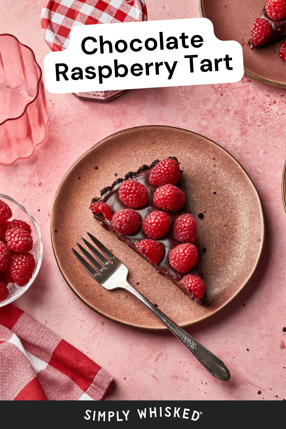 A slice of chocolate raspberry tart topped with fresh raspberries sits elegantly on a brown plate, with a fork beside it. Surrounding the decadent chocolate raspberry tart are more raspberries, a pink glass, and a jar with a red and white checked lid.