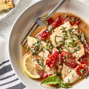 A bowl of fish fillet topped with chopped basil and cherry tomatoes in a light broth, garnished with a lemon slice, with a fork resting on the edge. Bread and a drink are partially visible nearby.