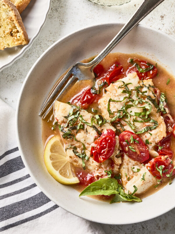 A bowl of fish fillet topped with chopped basil and cherry tomatoes in a light broth, garnished with a lemon slice, with a fork resting on the edge. Bread and a drink are partially visible nearby.