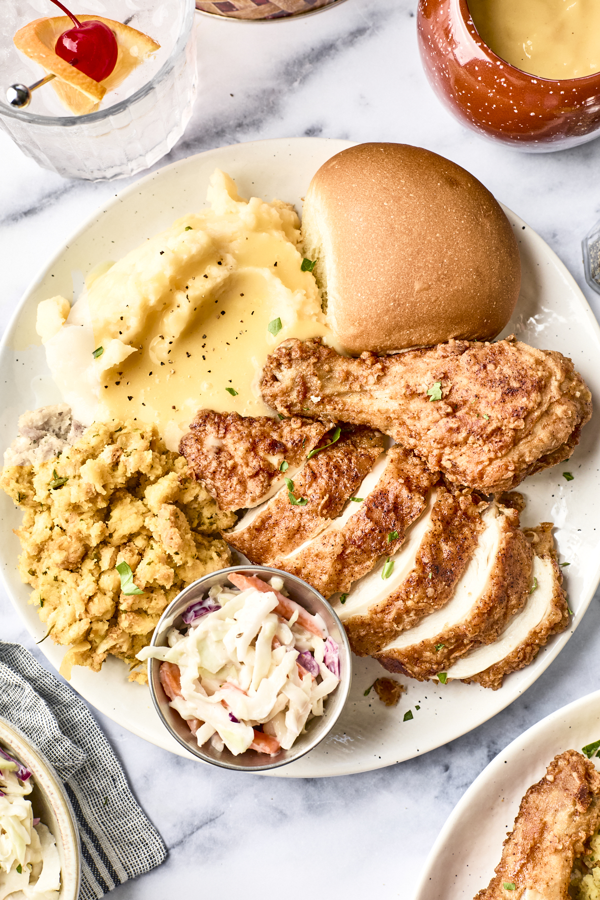 A plate of fried chicken, sliced chicken breast, mashed potatoes with gravy, stuffing, a dinner roll, and a small serving of coleslaw. Drinks and extra gravy are visible in the background.