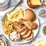 A plate with fried chicken, mashed potatoes with gravy, stuffing, coleslaw, and a bread roll is surrounded by bowls of coleslaw, chopped herbs, salt and pepper shakers, and a drink with a cherry garnish.