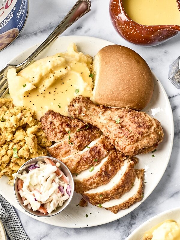 A plate with fried chicken, mashed potatoes with gravy, stuffing, coleslaw, and a bread roll is surrounded by bowls of coleslaw, chopped herbs, salt and pepper shakers, and a drink with a cherry garnish.