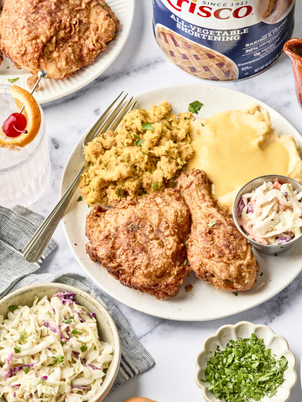 A plate with fried chicken, mashed potatoes with gravy, stuffing, and coleslaw sits on a table next to a bowl of extra coleslaw, a drink with an orange slice, fresh herbs, and a can of Crisco shortening.