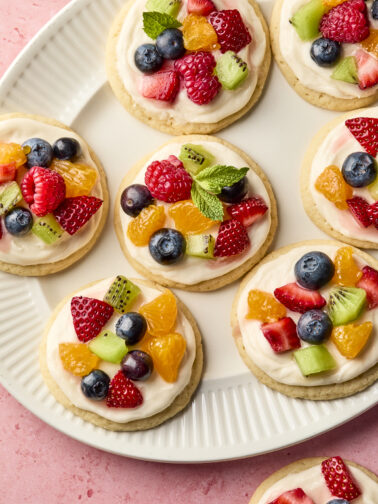 A plate of round sugar cookies topped with cream frosting and decorated with fresh berries, kiwi, and mint leaves, arranged on a white plate over a pink surface.
