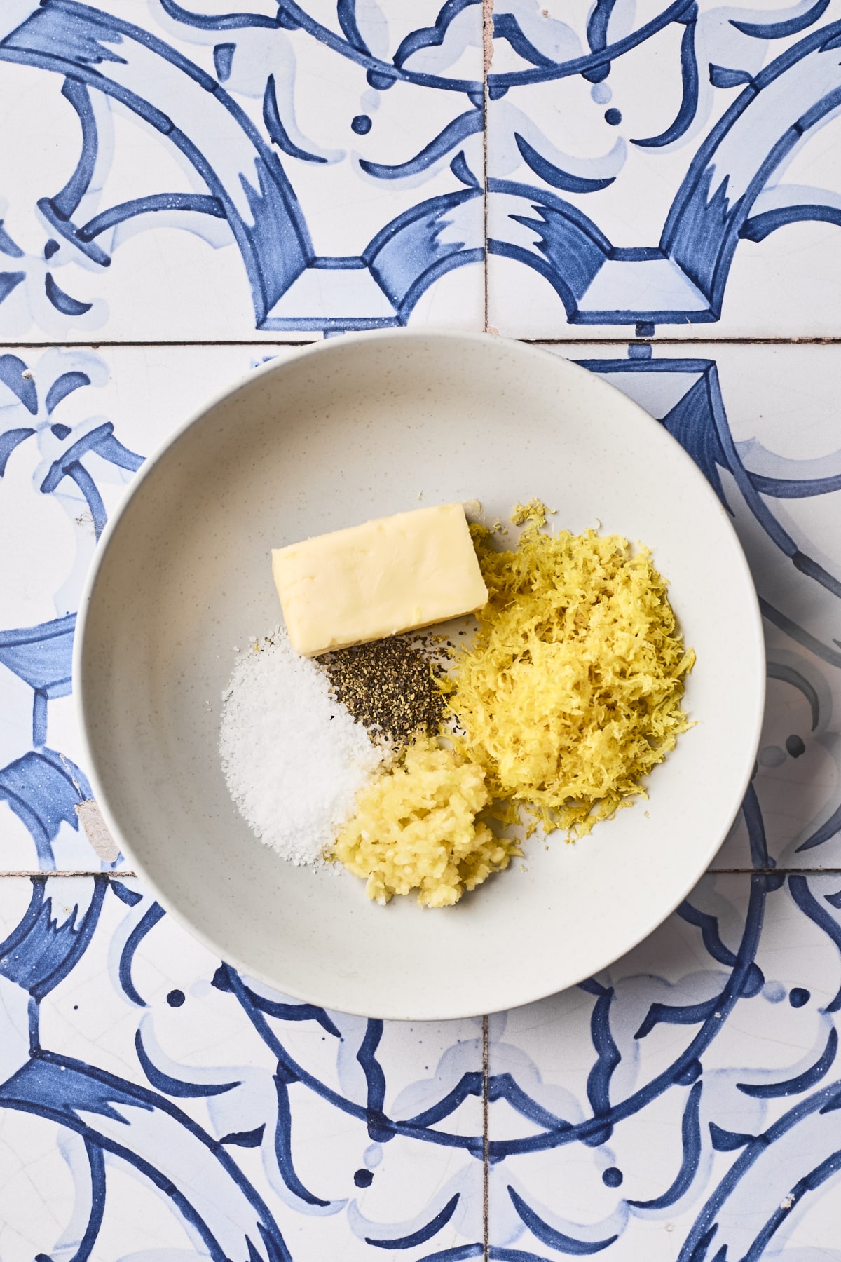 A white plate on a blue and white tiled surface holds a pat of butter, a pile of lemon zest, minced garlic, salt, and black pepper.