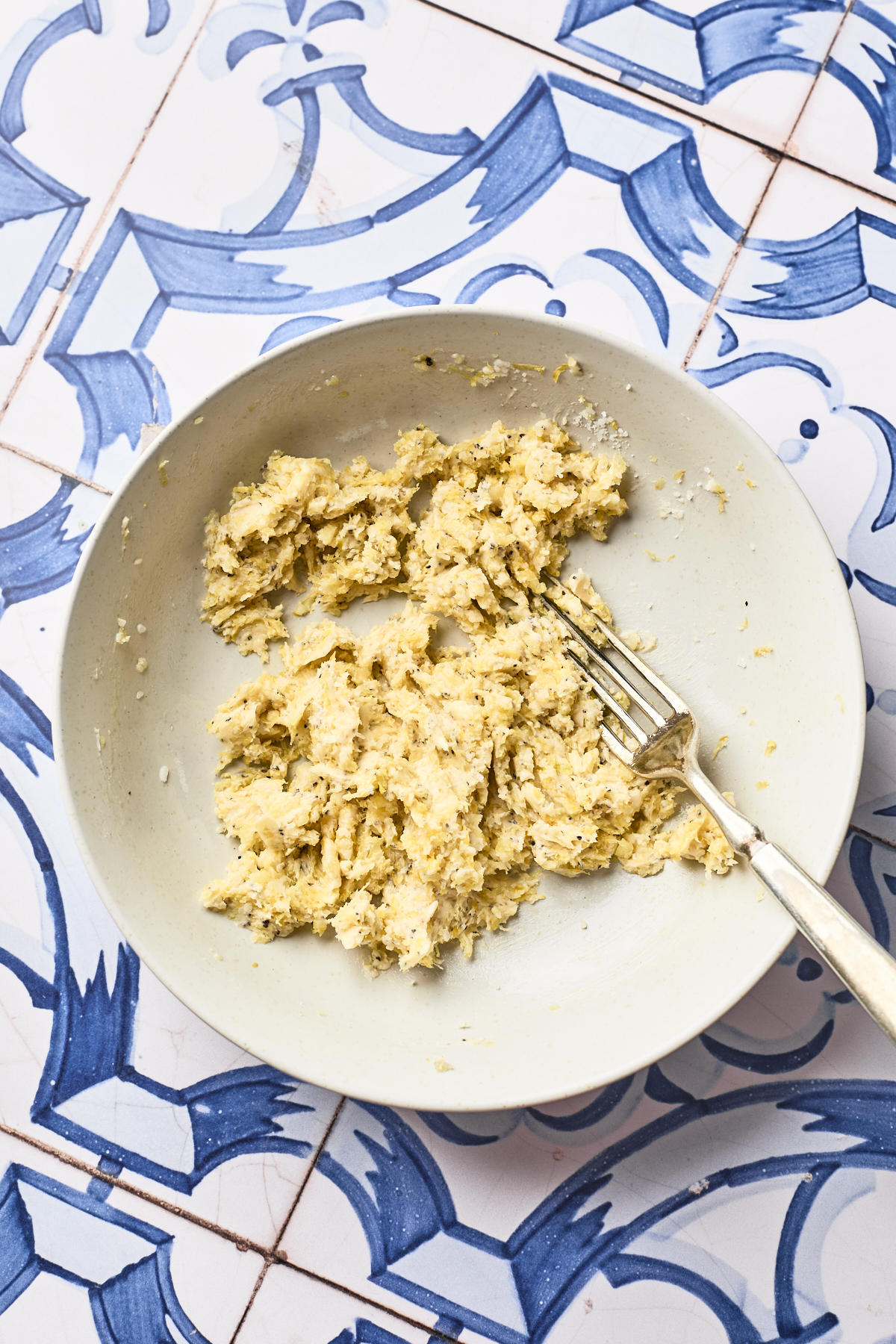 A white bowl with a fork and a mixture of shredded chicken and seasonings sits on a blue and white patterned tile surface.