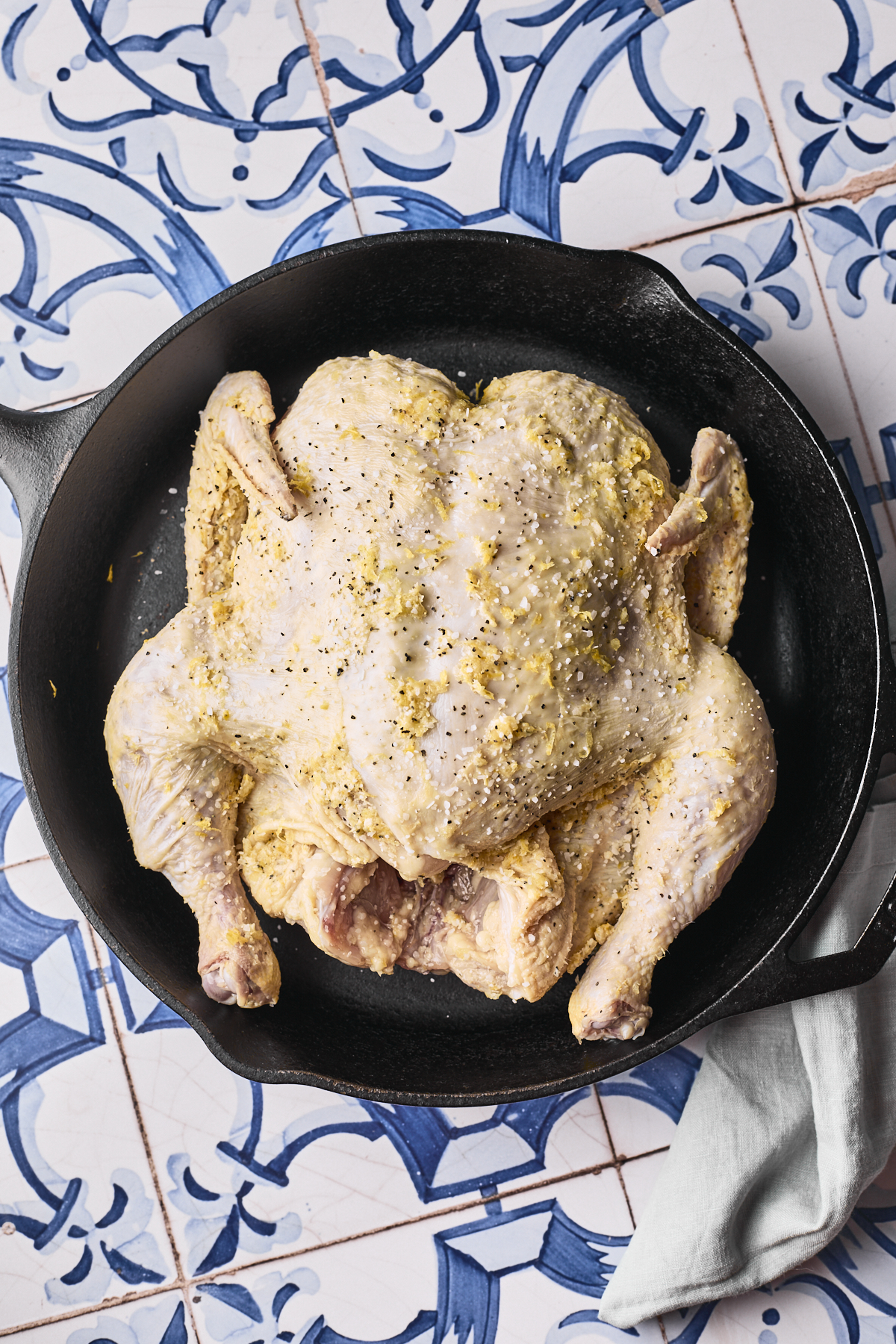 A whole raw chicken seasoned with herbs and pepper sits in a black cast iron skillet on a blue and white patterned tile surface, with a light gray cloth nearby.