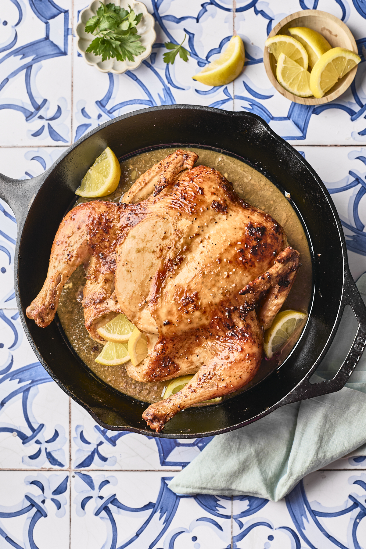 A roasted spatchcock chicken sits in a cast iron skillet with lemon wedges and juices, placed on a blue-patterned tile surface with lemon slices, parsley, and a green napkin nearby.