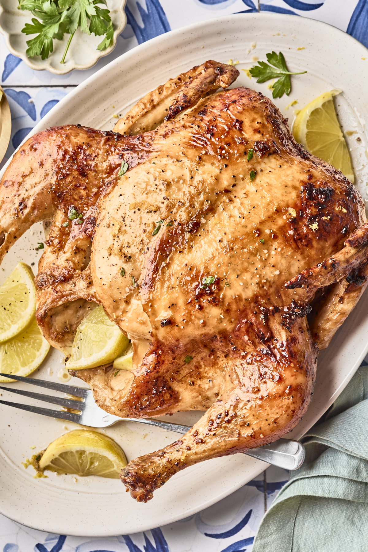 A roasted whole chicken garnished with chopped herbs, served on a white platter with lemon wedges. A fork rests on the platter, and a napkin and parsley are visible nearby.