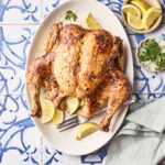 A whole roasted chicken on an oval platter, garnished with lemon wedges and parsley, set on a blue and white patterned table with a folded napkin, fork, extra lemon slices, and parsley nearby.