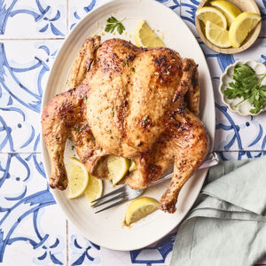 A whole roasted chicken on an oval platter, garnished with lemon wedges and parsley, set on a blue and white patterned table with a folded napkin, fork, extra lemon slices, and parsley nearby.