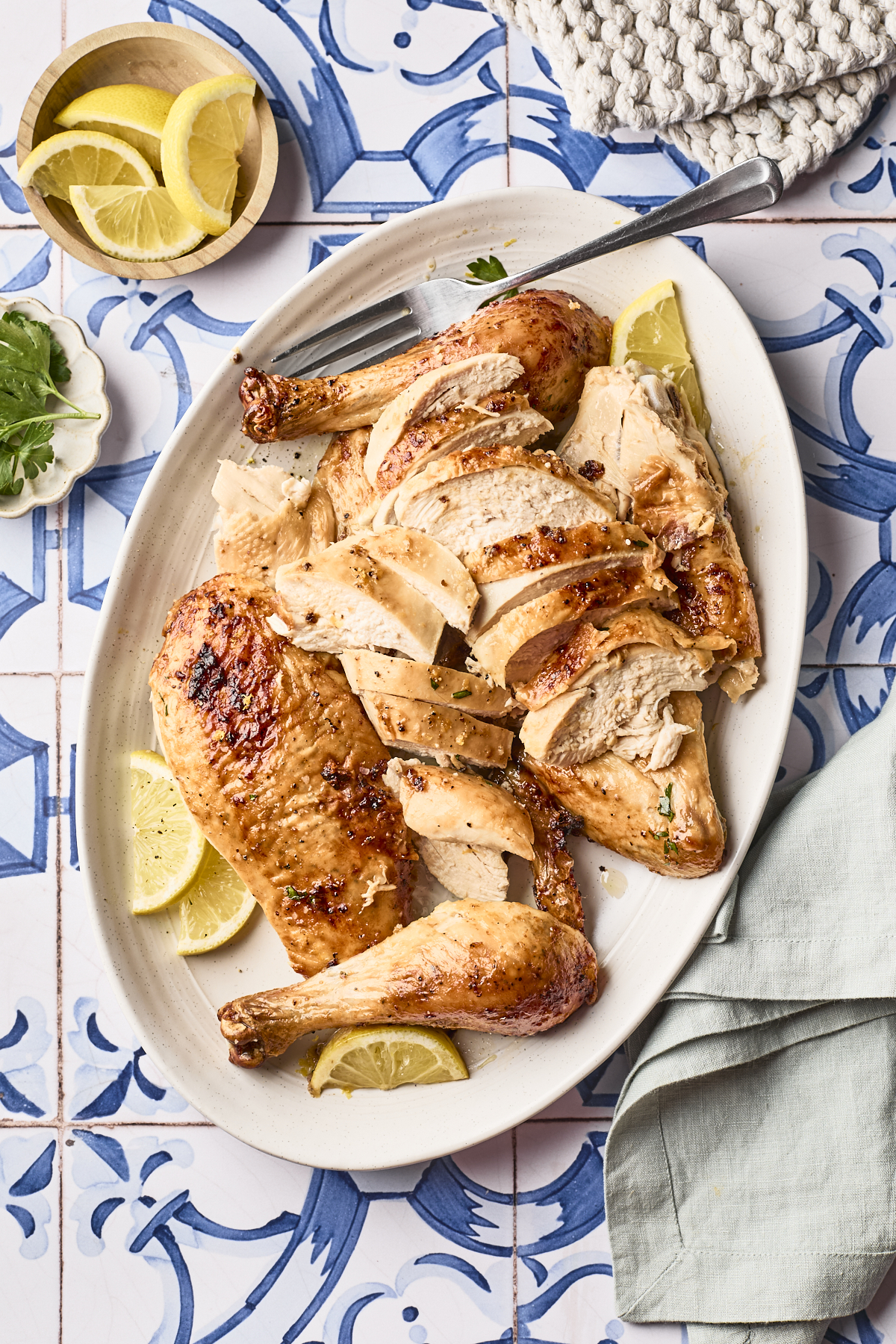 Sliced roasted chicken served on a white oval platter with lemon wedges, placed on a blue and white tiled surface. A small bowl of lemon slices, parsley, and a folded napkin are nearby.