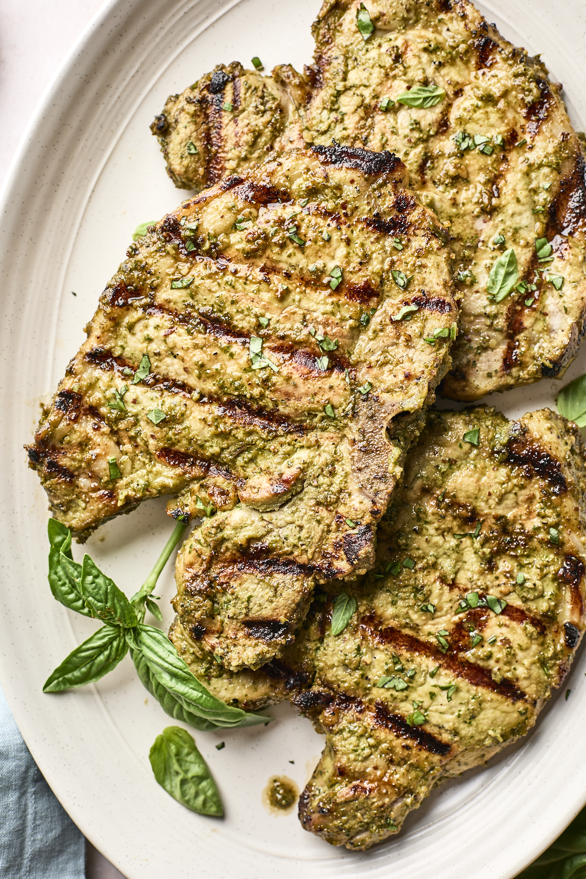Two grilled pork chops with visible grill marks, topped with chopped herbs, are served on a white oval plate. Fresh basil leaves are placed beside the chops for garnish.