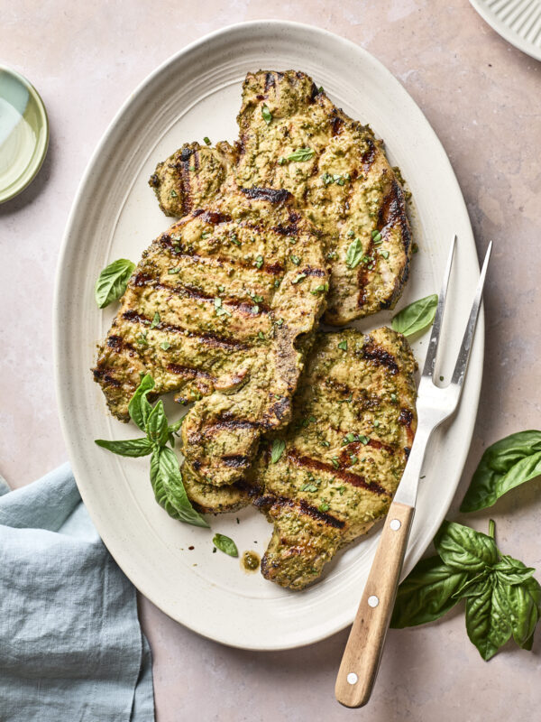 Grilled pork chops with visible grill marks on a white oval plate, garnished with fresh basil leaves. A meat fork rests beside the chops, and a light blue napkin is partially visible nearby.