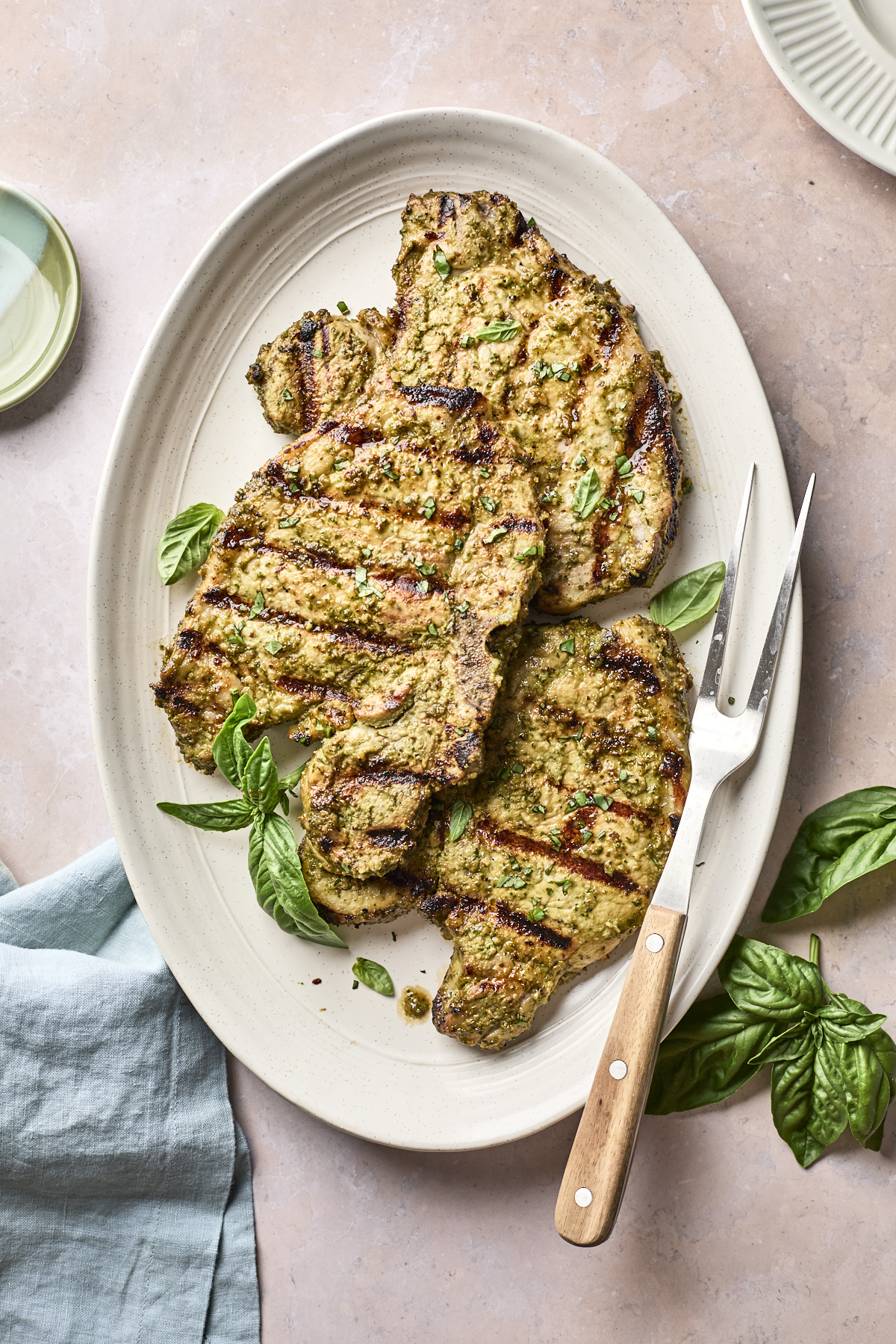 Grilled pork chops with visible grill marks on a white oval plate, garnished with fresh basil leaves. A meat fork rests beside the chops, and a light blue napkin is partially visible nearby.
