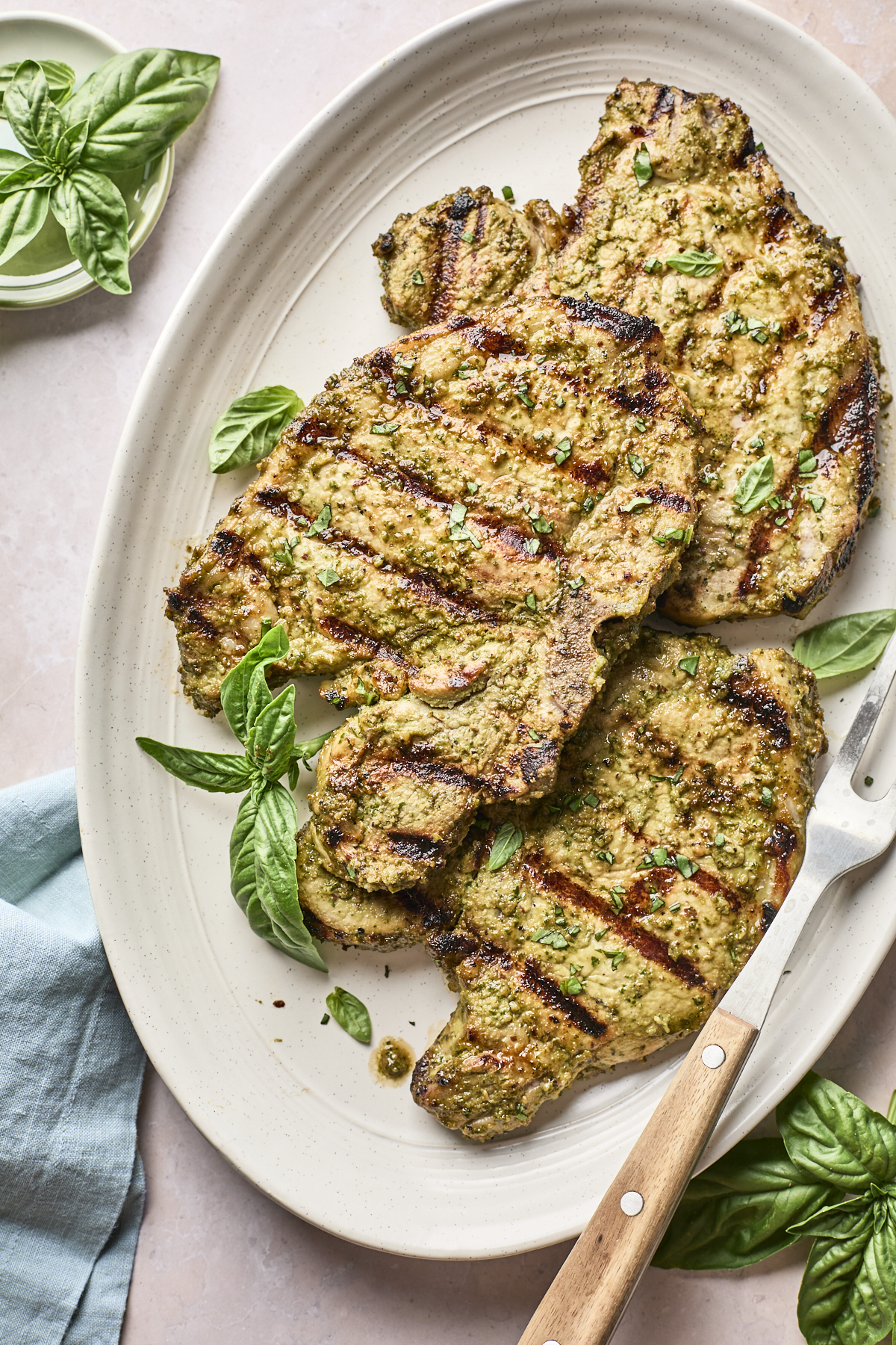 Grilled marinated chicken breasts with visible grill marks on an oval platter, garnished with fresh basil leaves. A fork and basil sprigs are nearby, and a light green napkin is placed on the side.