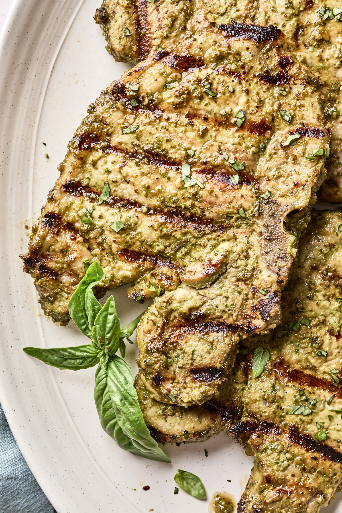 Grilled pork chops with visible char marks, garnished with chopped herbs and fresh basil leaves, served on a white plate.