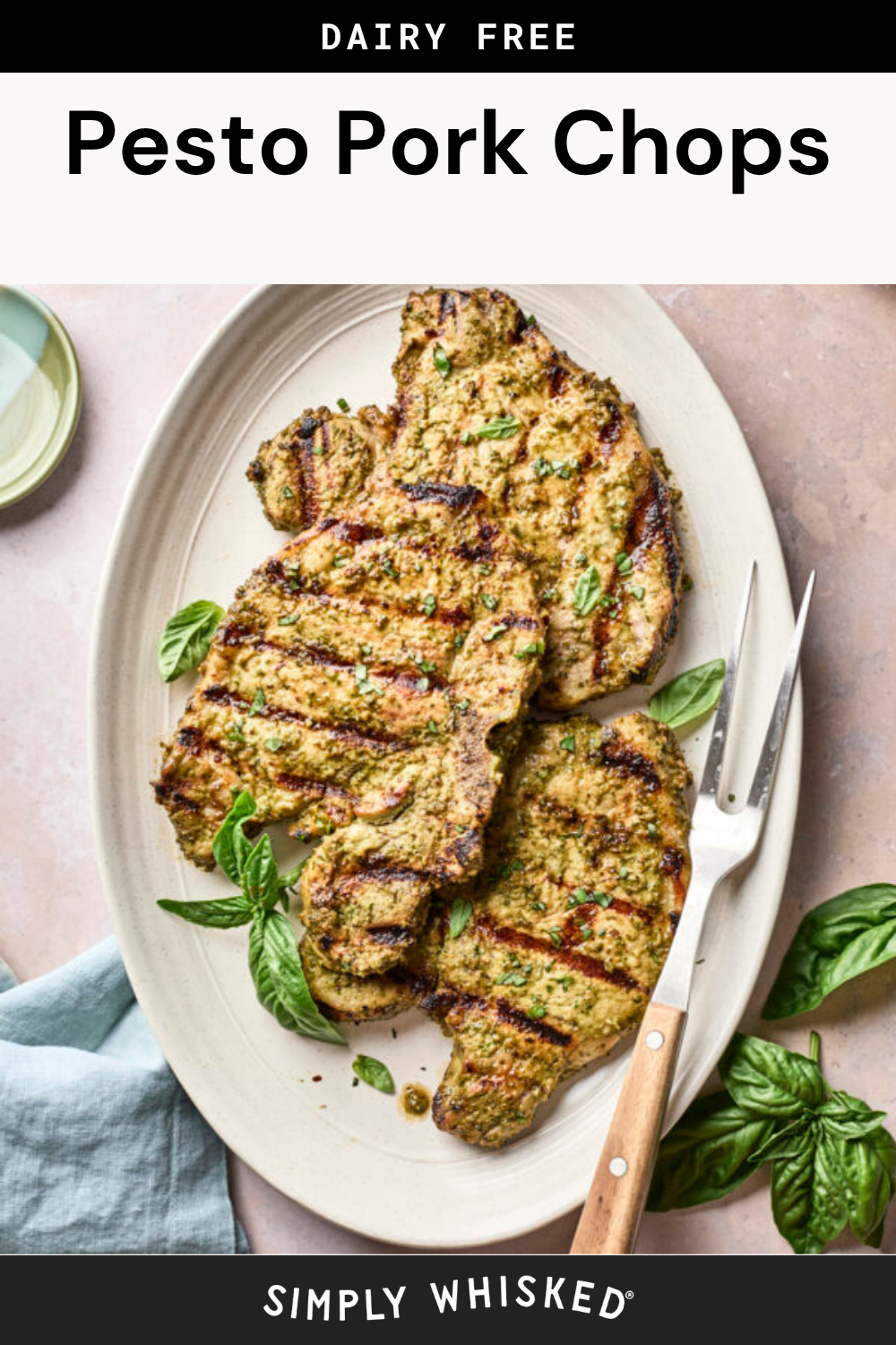 Grilled pesto pork chops coated with vibrant sauce are served on an oval plate, garnished with fresh basil leaves. A serving fork rests beside the chops. The image is labeled “Dairy Free Pesto Pork Chops.”.