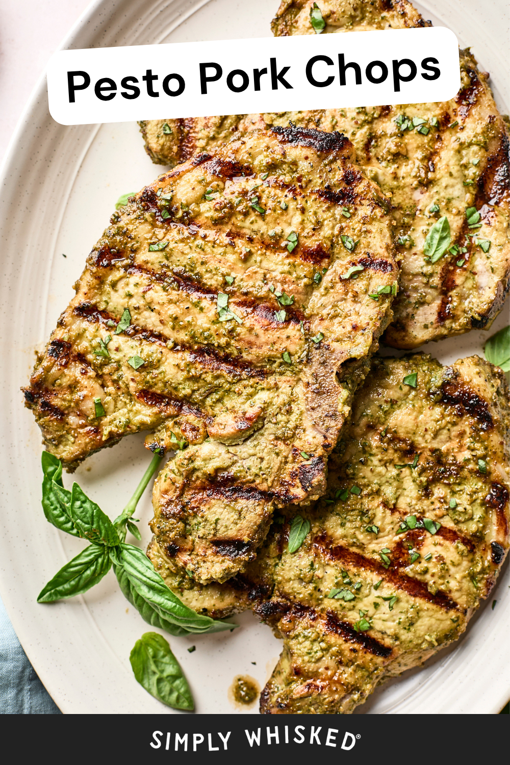 Grilled pesto pork chops coated with pesto, garnished with fresh herbs, and served on a light plate with basil leaves. Text reads Pesto Pork Chops at the top and Simply Whisked at the bottom.
