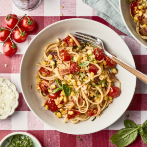 A bowl of pasta with cherry tomatoes, corn, basil, and cheese sits on a red-and-white checkered tablecloth, surrounded by fresh ingredients and a fork.