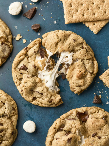 A chocolate chip cookie broken in half with gooey melted marshmallow inside, surrounded by whole cookies, chocolate chunks, mini marshmallows, and graham crackers on a blue surface.