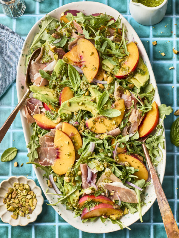 A platter of salad with arugula, sliced peaches, avocado, prosciutto, red onion, fresh basil, and a drizzle of green pesto, served on a turquoise tiled surface with utensils and a small bowl of pistachios nearby.