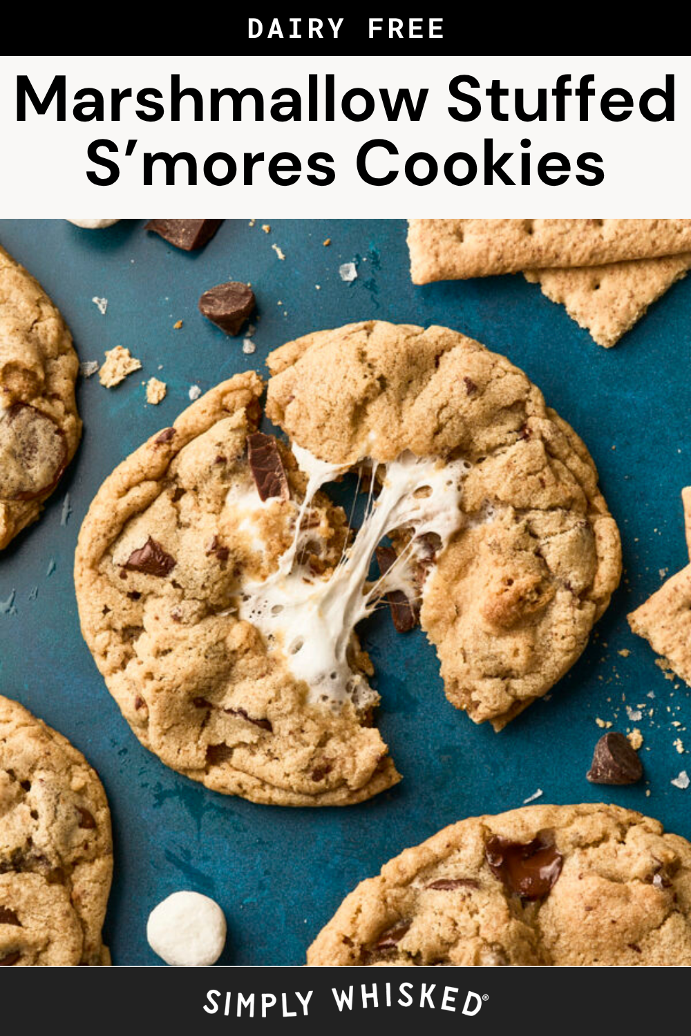 A close-up of dairy-free s’mores cookies on a blue background. The center cookie is broken in half, revealing gooey marshmallow and chocolate filling. Chocolate chunks and cookie crumbs are scattered around.