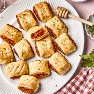 A white plate with several golden-brown sausage rolls, one cut open to show the filling. A honey dipper rests nearby, with a bowl of honey, green herbs, and a red-checked napkin beside the plate.