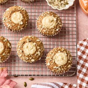 Pumpkin muffins topped with oats and creamy filling on a cooling rack, next to a bowl of oats, a checked kitchen towel, and a pumpkin-shaped container on a pink surface.