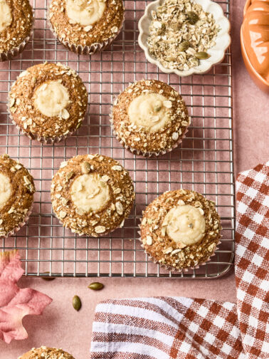 Pumpkin muffins topped with oats and creamy filling on a cooling rack, next to a bowl of oats, a checked kitchen towel, and a pumpkin-shaped container on a pink surface.