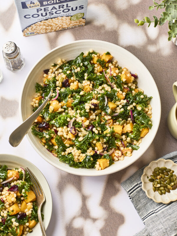 A bowl of pearl couscous salad with kale, roasted butternut squash, red onions, cranberries, and pumpkin seeds. A fork, a small plate of seeds, a box of couscous, and a plant are nearby on a patterned tablecloth.