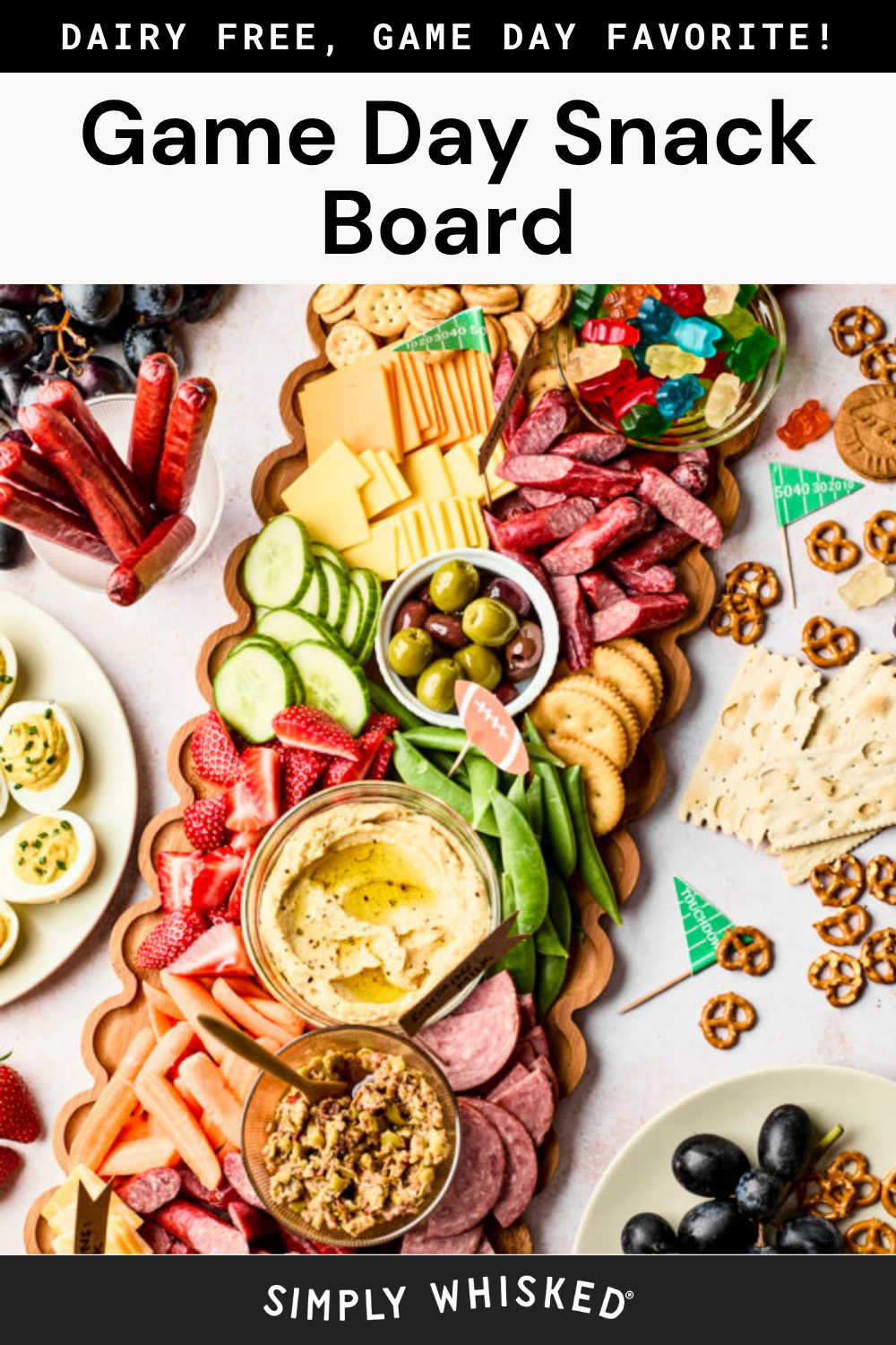 A colorful snack board with dairy-free cheese, hummus, veggies, olives, meats, crackers, gummies, and grapes, arranged for a game day party. Text reads: DAIRY FREE, GAME DAY FAVORITE! Game Day Snack Board..