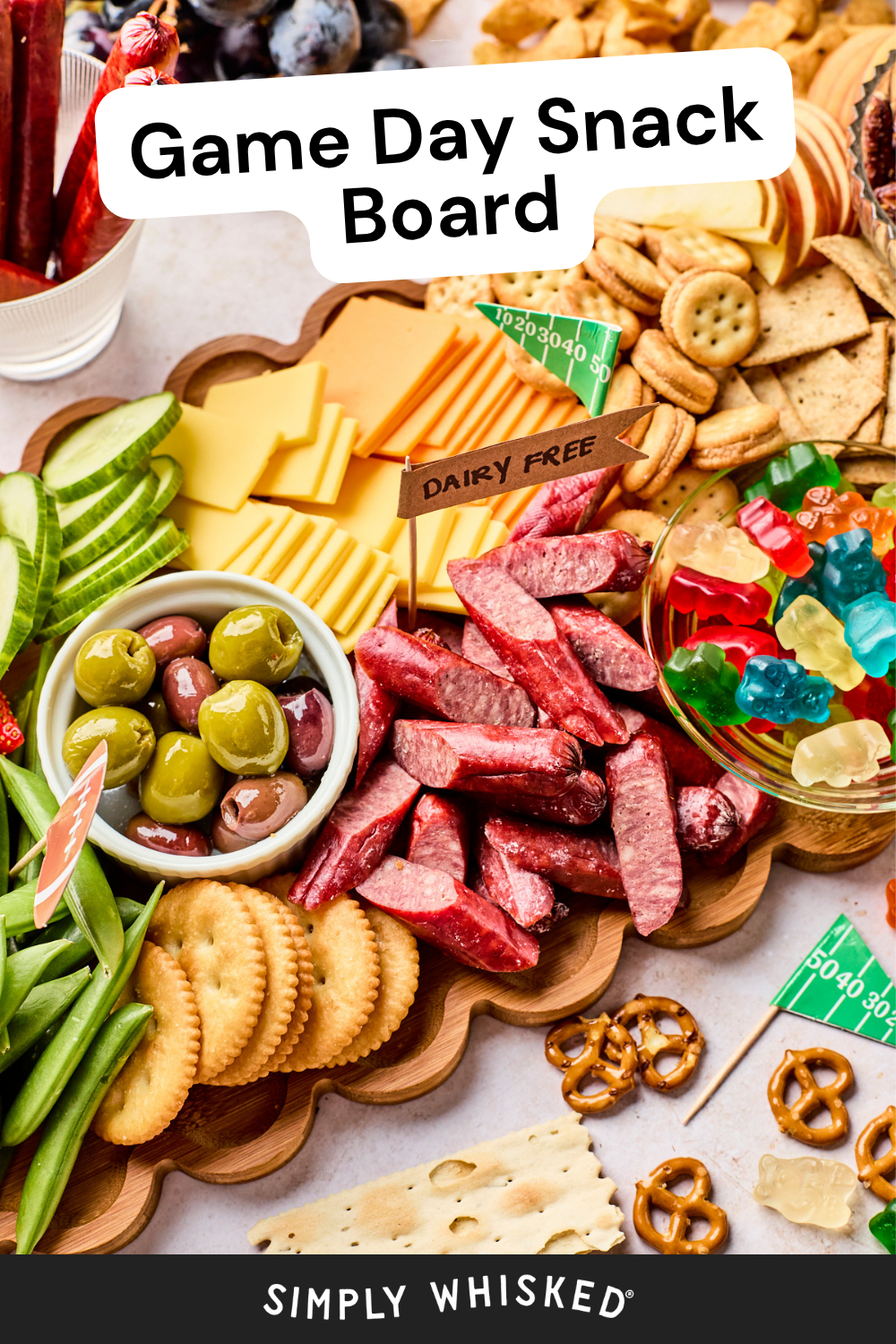 A game day snack board with crackers, cheese, olives, sliced meats, pretzels, cucumber, sugar snap peas, and gummy bears on a wooden platter. Sign reads Dairy Free. Text: Game Day Snack Board..