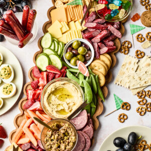 A wooden platter filled with assorted cheeses, crackers, meats, olives, hummus, cucumber slices, strawberries, and snap peas, surrounded by plates of grapes, deviled eggs, pretzels, and other snacks.