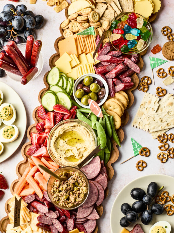 A wooden platter filled with assorted cheeses, crackers, meats, olives, hummus, cucumber slices, strawberries, and snap peas, surrounded by plates of grapes, deviled eggs, pretzels, and other snacks.