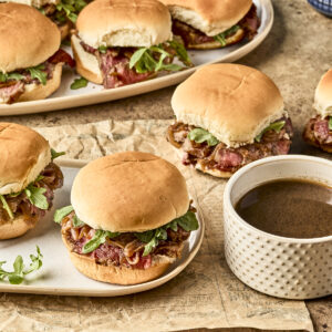Plates of sandwiches with roast beef, greens, and onions on soft buns, served on a rustic surface with a cup of brown dipping sauce nearby.