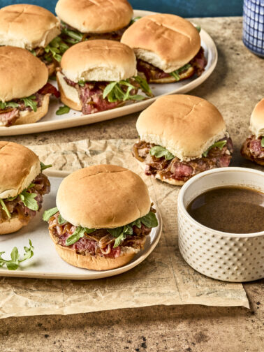 Plates of sandwiches with roast beef, greens, and onions on soft buns, served on a rustic surface with a cup of brown dipping sauce nearby.