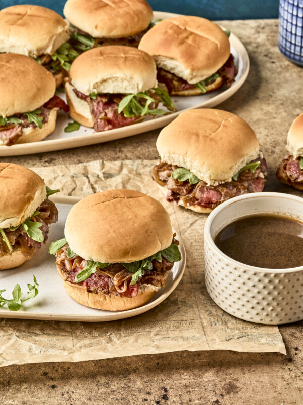 Plates of sandwiches with roast beef, greens, and onions on soft buns, served on a rustic surface with a cup of brown dipping sauce nearby.