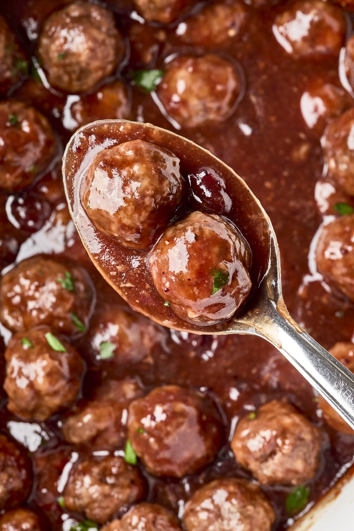 A spoon holding two glazed meatballs in a rich, dark red sauce, with more meatballs visible in the saucy background.