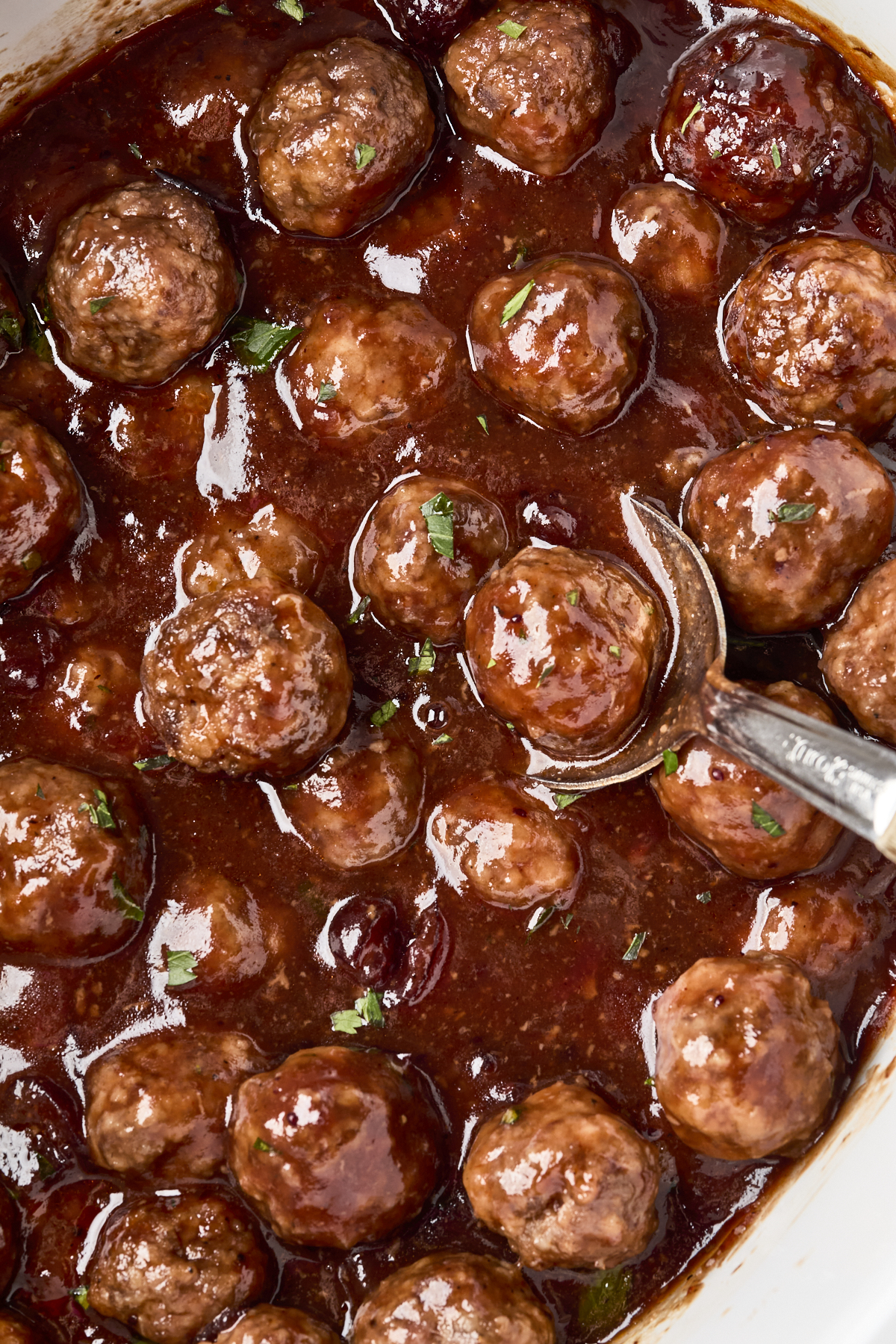 A close-up of meatballs coated in a thick, glossy reddish-brown sauce, garnished with small bits of green herbs, with a metal spoon partially visible among the meatballs.