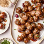 A large oval platter filled with glazed meatballs garnished with herbs, with some on toothpicks. Cranberries and fresh herbs are scattered around, and a small plate holds extra meatballs. A bundle of toothpicks and a napkin are nearby.