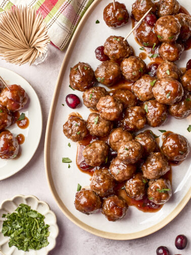 A large oval platter filled with glazed meatballs garnished with herbs, with some on toothpicks. Cranberries and fresh herbs are scattered around, and a small plate holds extra meatballs. A bundle of toothpicks and a napkin are nearby.