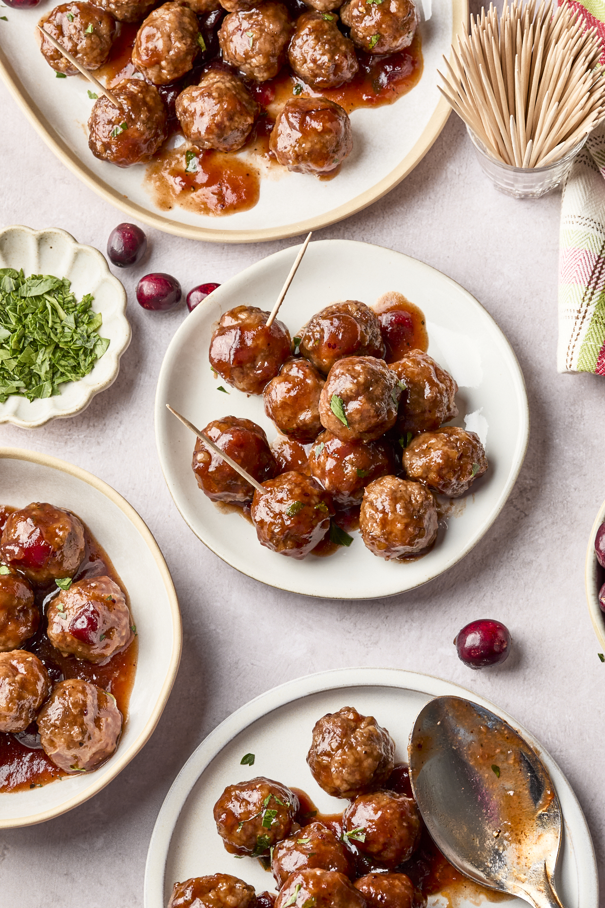 Four plates of glazed cocktail meatballs garnished with herbs, some with toothpicks inserted. A dish of chopped parsley, scattered cranberries, and a container of toothpicks are also visible on a light surface.