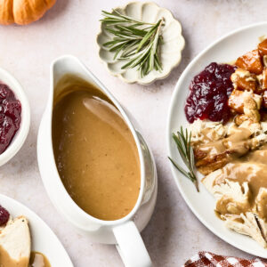 A Thanksgiving meal with gravy in a white boat, sliced turkey with gravy and cranberry sauce on a plate, stuffing, mashed potatoes, a sprig of rosemary, a small pumpkin, and a red checkered napkin.