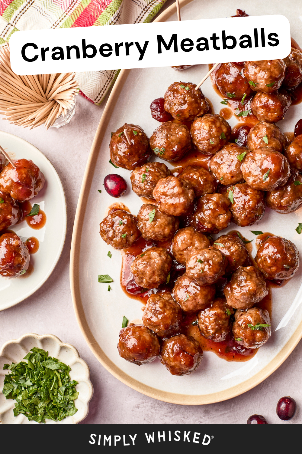 A platter of glazed cranberry meatballs garnished with herbs, with a small serving on a plate nearby. Fresh cranberries and a dish of chopped herbs are placed alongside. Text reads Cranberry Meatballs..
