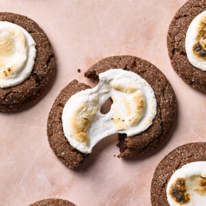 Chocolate cookies topped with toasted marshmallows are arranged on a light pink surface. One cookie in the center is broken in half, showing its soft texture and gooey marshmallow topping.