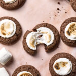 Chocolate cookies topped with toasted marshmallows are arranged on a pink surface. One cookie is broken in half, revealing a gooey marshmallow center. Extra marshmallows and cookie crumbs are also visible.
