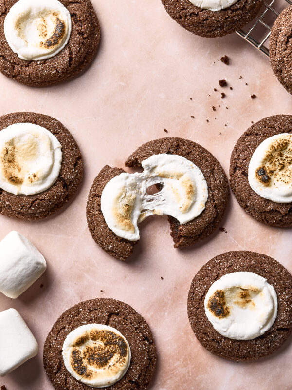 Chocolate cookies topped with toasted marshmallows are arranged on a pink surface. One cookie is broken in half, revealing a gooey marshmallow center. Extra marshmallows and cookie crumbs are also visible.